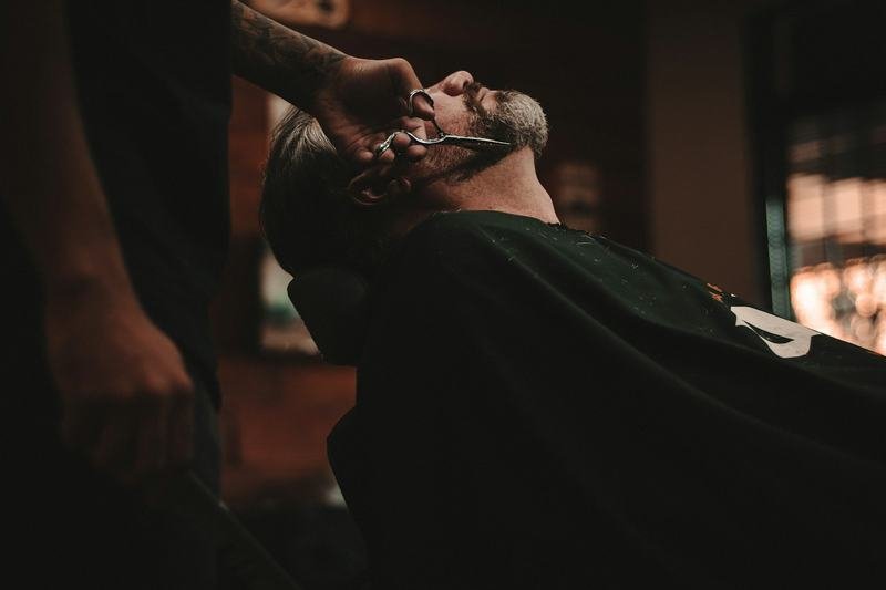 Barbeiro fazendo a barba de um cliente reclinado na cadeira, em luz baixa