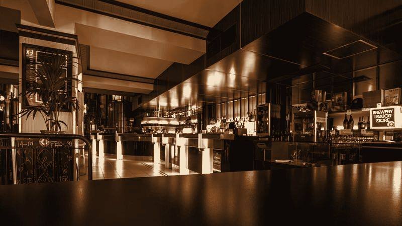 Salão do restaurante em luz baixa, com balcão iluminado ao fundo